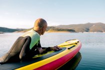 Side view of male in wetsuit lying on paddle board and swimming on lake surface while practicing water sport in summer day — Stock Photo