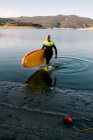 Мужчина серфер в гидрокостюме ходьба с желтой доской SUP и весло в песчаном пляже рядом с морской водой — стоковое фото