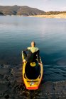 Vista posterior de un hombre irreconocible y reflexivo en traje de neopreno sentado en el tablero de paddle mientras se prepara para remar en el agua del lago - foto de stock