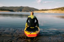 Vista posterior de un hombre irreconocible y reflexivo en traje de neopreno sentado en el tablero de paddle mientras se prepara para remar en el agua del lago - foto de stock