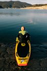 Задний вид неузнаваемого вдумчивого мужчины в гидрокостюме, сидящего на веслах во время подготовки к гребле в озере — стоковое фото