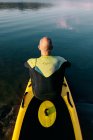 Visão traseira do macho pensativo irreconhecível em roupa de mergulho sentado na prancha de remo enquanto se prepara para remar na água do lago — Fotografia de Stock