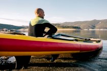 Назад погляд спокійний чоловік-серфер з веслом, що сидить на борту позашляховика на піщаному пляжі біля спокійного моря і насолоджується видом. — стокове фото