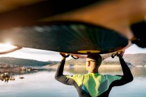 Rückenansicht eines bis zur Unkenntlichkeit gereiften männlichen Surfers, der abends mit dem Paddelbrett über dem Kopf in der Nähe des Meeres steht und wegschaut — Stockfoto