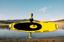 Side view of cropped unrecognizable male surfer in wetsuit standing with yellow SUP board and paddle in sea water — Stock Photo