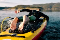 Вид ззаду на невпізнаваного босоніж чоловіка в гідрокостюмі лежить на дошці весла і плаває на поверхні озера, займаючись водними видами спорту в літній день — стокове фото