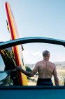 Rückenansicht des aktiven Mannes mittleren Alters ohne Hemd, der das Paddelbrett aus dem am Seeufer geparkten Auto in hügeliger Natur nimmt, während er sich auf die Wassersportübungen im Sommer vorbereitet — Stockfoto