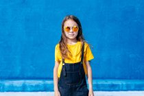 Selbstbewusster Teenager steht mit trendiger Sonnenbrille vor blauem Hintergrund und blickt in die Kamera — Stockfoto