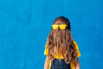 Anónimo fresco adolescente usando gafas de sol amarillas en el pelo largo cubriendo la cara sobre fondo azul - foto de stock