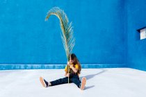 Підліток сидить з величезним пальмовим листом на фоні синьої стіни — стокове фото