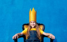 Зміст підлітка в золотій паперовій короні, що сидить на троні, як король на синьому фоні, дивлячись на камеру — стокове фото