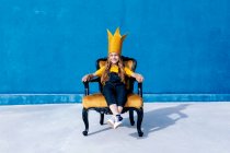 Contenido adolescente en la corona de papel dorado sentado en el trono como rey sobre fondo azul mirando a la cámara - foto de stock