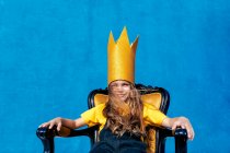 Содержание подросток в золотой бумажной короне сидит на троне, как король на синем фоне, глядя в камеру — стоковое фото