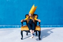 Content teenager in golden paper crown sitting on throne like king on blue background looking away — Stock Photo