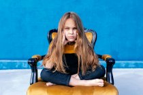 Sad teenager with long hair and in hipster clothes sitting on chair with legs crossed while looking at camera on blue background — Stock Photo