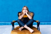 Trauriger Teenager mit langen Haaren und in Hipster-Klamotten, der mit gekreuzten Beinen auf einem Stuhl sitzt und vor blauem Hintergrund wegschaut — Stockfoto