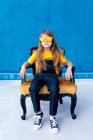 Thoughtful teen hipster sitting on chair with trendy yellow sunglasses on blue background looking at camera — Stock Photo