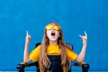 Ausdrucksstarker Teenager mit langen Haaren und trendiger gelber Sonnenbrille mit Rockzeichen und Schreien auf blauem Hintergrund — Stockfoto