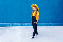 Прохолодний вдумливий підліток хіпстер в комбінезоні і жовтий капелюх, що йде руками в кишенях на тлі синьої стіни — стокове фото