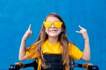 Expressive teenager with long hair and in trendy yellow sunglasses showing rock sign and screaming on blue background — Stock Photo