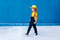 Cool hipster adolescente reflexivo en overol y sombrero amarillo caminando con las manos en los bolsillos en el fondo de la pared azul - foto de stock