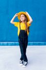 Cool hipster adolescente alegre en overoles y sombrero amarillo de pie mirando hacia otro lado en el fondo de la pared azul - foto de stock
