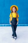 Coole, fröhliche Teenager-Hipster in Overalls und gelbem Hut, die Hände in Taschen auf blauem Wandhintergrund und in die Kamera blickend — Stockfoto