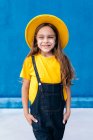 Cool alegre adolescente hipster em macacão e chapéu amarelo de pé com as mãos em bolsos no fundo da parede azul olhando para a câmera — Fotografia de Stock