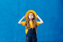 Cooler nachdenklicher Teenager-Hipster in Overalls und gelbem Hut, der mit geschlossenen Augen vor dem Hintergrund der blauen Wand steht — Stockfoto