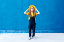 Cool hipster adolescente reflexivo en overol y sombrero amarillo de pie con los ojos cerrados en el fondo de la pared azul - foto de stock