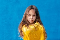 Grave adolescente con i capelli lunghi avvolto in mantello giallo in piedi su sfondo blu e guardando la fotocamera — Foto stock