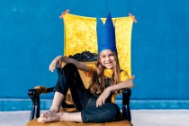 Contenido adolescente en la corona de papel dorado sentado en el trono como rey sobre fondo azul mirando a la cámara - foto de stock