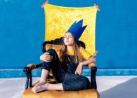 Nachdenklicher Teenager in goldener Papierkrone, der wie ein König auf blauem Hintergrund mit geschlossenen Augen auf dem Thron sitzt — Stockfoto