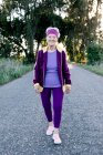 Gusto jogger femenina mayor sonriendo y caminando por el camino de asfalto durante el entrenamiento de fitness en la mañana de verano en el campo - foto de stock