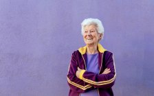 Candid atleta anziana donna in abiti sportivi guardando lontano con le braccia incrociate su sfondo viola — Foto stock