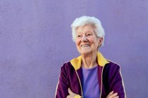 Candid atleta anziana donna in abiti sportivi guardando lontano con le braccia incrociate su sfondo viola — Foto stock