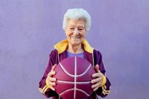 Sonriente jugadora de baloncesto anciana con pelo gris en ropa deportiva mirando a la cámara sobre fondo violeta - foto de stock