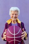Lächelnde ältere Basketballspielerin mit grauen Haaren in Sportkleidung blickt in die Kamera auf violettem Hintergrund — Stockfoto