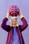 Sorridente anziano giocatore di basket femminile con i capelli grigi in abiti sportivi guardando la fotocamera su sfondo viola — Foto stock