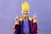 Zufriedene Seniorinnen in Sportbekleidung und Papierkrone zeigen mit den Fingern nach oben, während sie in die Kamera auf violettem Hintergrund schauen — Stockfoto