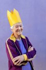 Confident elderly female athlete in sportswear and decorative crown looking at camera with folded arms on purple background — Stock Photo