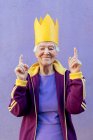 Conteúdo Atleta feminina sênior em sportswear e coroa de papel apontando para cima com os dedos enquanto olha para a câmera no fundo violeta — Fotografia de Stock