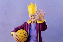 Весела старша жінка-спортсменка в спортивному одязі з баскетболом дивиться на камеру, демонструючи жест управління на фіолетовому фоні — стокове фото