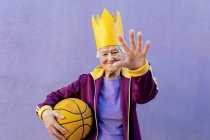 Alegre atleta senior en ropa deportiva con baloncesto mirando a la cámara mientras demuestra un gesto de control sobre fondo púrpura - foto de stock