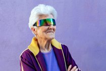 Alegre atleta anciana con los brazos cruzados y pelo gris en ropa deportiva y venda en el fondo violeta - foto de stock