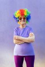 Содержание старшей спортсменки в современных солнцезащитных очках и ярком парике, стоящем со сложенными руками на фиолетовом фоне — стоковое фото