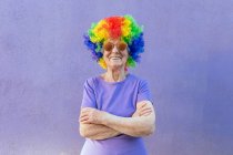 Contenuto atleta donna anziana in occhiali da sole moderni e parrucca luminosa in piedi con le braccia piegate su sfondo viola — Foto stock
