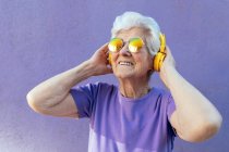 Fröhliche Seniorin in T-Shirt und moderner Sonnenbrille hört Song aus drahtlosem Headset auf lila Hintergrund — Stockfoto