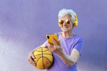 Весела літня жінка в сучасних сонцезахисних окулярах та навушниках слухати музику під час серфінгу в Інтернеті на мобільному телефоні на фіолетовому фоні — стокове фото