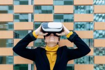 Female in jacket and turtleneck using VR goggles near building windows in daylight — Stock Photo